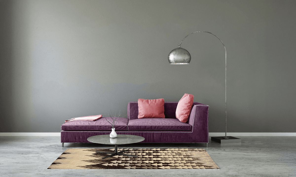 Sala de estar moderna con un sofá morado, cojines rosas y una alfombra estampada sobre un suelo de concreto.
